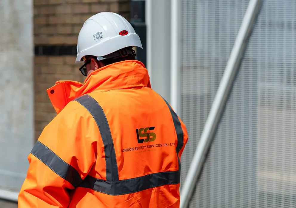 A person wearing a white hard hat and orange high-visibility jacket with reflective stripes and "London Security Services (UK) Ltd" logo stands outside a building.