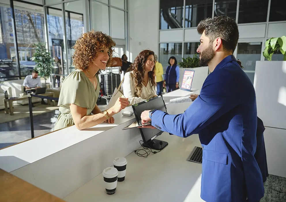 A woman at a reception desk smiles while handing a document to a man in a blue suit. Two coffee cups and a laptop are on the counter in a bright, modern lobby.