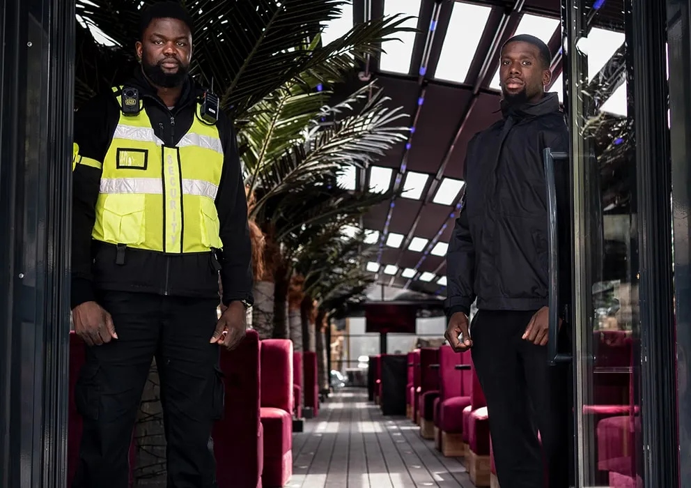 Two security guards stand at the entrance of a modern venue with palm trees and red seating. They appear serious and attentive.