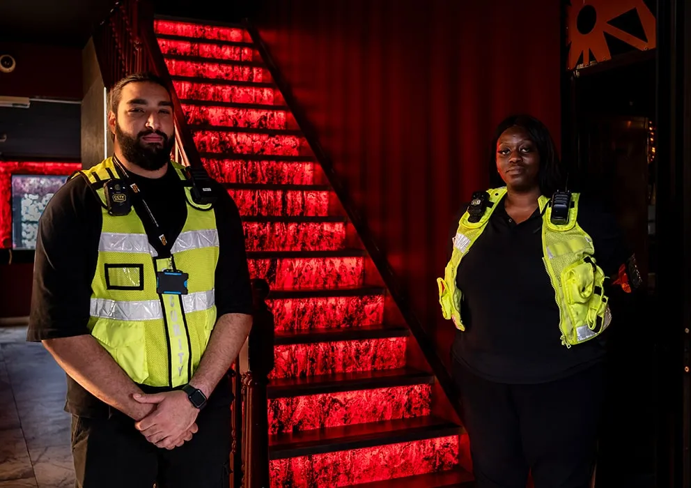 Two security staff in fluorescent vests stand confidently in a dimly lit space. Behind them, a staircase glows with vivid red lights, adding a dramatic effect.