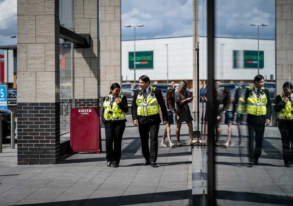 Two security officers in yellow vests walk by a glass building, their reflections visible. Behind them, people stand near a Costa Coffee sign. Sunny day.
