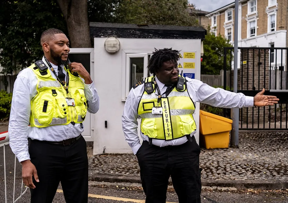 Two security guards stand outside a small building, wearing reflective vests and white shirts. One gestures to the right, indicating direction.